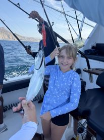 A young girl proudly displaying her catch, a small fish, while fishing on a boat in a scenic coastal environment.