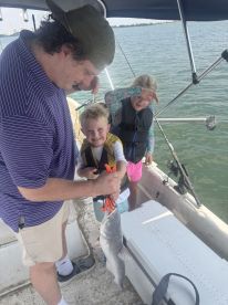 A young boy joyfully holds a caught fish with the help of an adult, while a girl looks on, all aboard a boat in a sunny fishing environment.