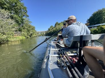 Two anglers navigating a scenic river in a fishing boat, showcasing a relaxed fishing environment with multiple rods visible.