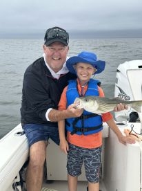 A young boy proudly displays a striped bass while fishing with an adult on a boat, showcasing a memorable fishing experience.