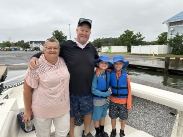 A family poses on the boat, ready for a fishing adventure, with two young boys in life jackets and their parents smiling at the dock.