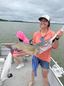 A smiling angler proudly displaying a large Catfish while fishing on a boat, with a scenic lake backdrop.