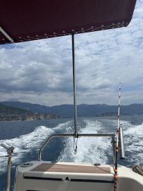 A scenic view from the back of a boat cruising through calm waters, showcasing the mountainous coastline under a partly cloudy sky.