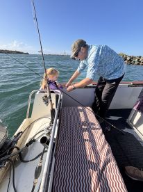 A young girl is being assisted by an adult while fishing on a boat, showcasing a hands-on learning experience in a scenic waterway.