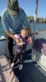 A young girl joyfully holds a small fish with the help of an adult on a fishing boat, showcasing a memorable fishing experience.