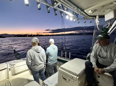 Three anglers aboard a boat during twilight, actively fishing as the sun sets over the water.