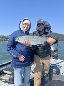 Two anglers proudly displaying a large Silver Salmon on a fishing boat, enjoying a sunny day on the water.