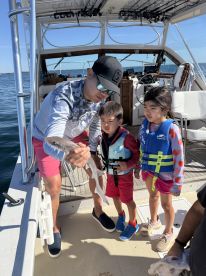 A young boy and girl, both wearing life jackets, watch excitedly as an adult holds a small shark on a fishing boat, showcasing a family fishing experience.