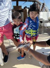 Two young children on a boat excitedly touching a small shark, showcasing a fun fishing experience.