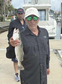 An angler proudly displaying a Speckled Trout caught during a fishing trip, with a boat docked in the background.
