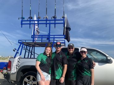 A group of four anglers posing in front of a fishing truck with a rod holder setup, showcasing their fishing gear at a beach location.