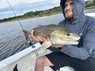 An angler proudly displaying a large Redfish while fishing from a boat in a scenic waterway.