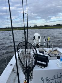 View of the boat's interior with fishing rods and a net, set against a scenic backdrop of water and greenery.