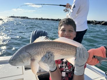 A young angler proudly displaying a large Redfish while fishing on a sunny day, with another fisherman in the background.