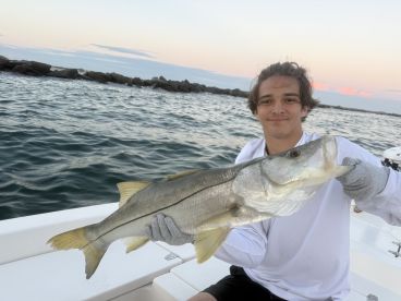 A young angler proudly displaying a large Snook while fishing from a boat in coastal waters during sunset.