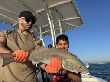 A young angler and an adult proudly displaying a large Redfish on a sunny day out on the water, showcasing a successful fishing trip.