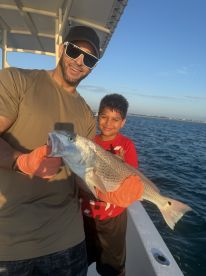A proud angler and a young boy display a large Snapper caught during a fishing trip, showcasing a beautiful day on the water.