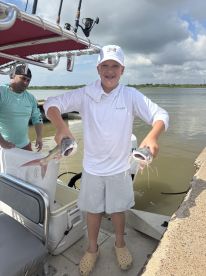 A young angler proudly displaying two Catfish while standing on a boat dock, showcasing a successful fishing trip.