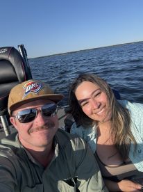 Two anglers enjoying a sunny day on the water, seated inside a fishing boat, with a scenic backdrop of the lake.