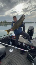 An angler proudly displaying a large Lake Trout while fishing on a calm lake, showcasing a successful day on the water.