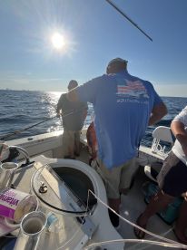 Anglers actively fishing on a boat in open water, with a sunny backdrop and visible fishing gear.
