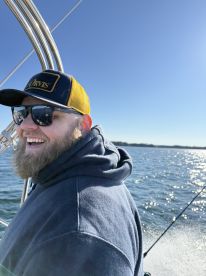 A smiling angler enjoying a sunny day on the water, showcasing the boat's exterior and the sparkling water in the background.
