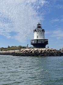 A scenic view of a lighthouse with visitors on the observation deck, set against a clear blue sky and calm waters.