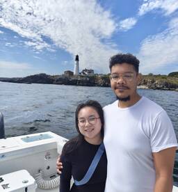A couple poses together on a boat with a scenic lighthouse in the background, enjoying a day on the water.