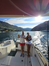 Two friends enjoying a sunny day on a boat in a scenic coastal area, with mountains in the background.