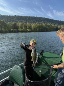 A young angler proudly displaying a caught Bass while fishing on a serene lake surrounded by lush greenery.