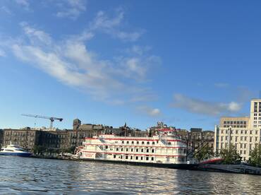 The image showcases the exterior of the 'Georgia Queen,' a riverboat, docked along the waterfront with a clear blue sky in the background.
