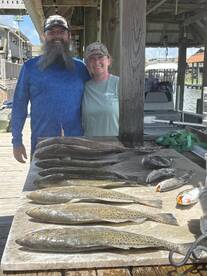 Two anglers proudly posing with a variety of fish, including Speckled Trout and Redfish, displayed on a cleaning table at a dock.