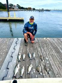 An angler kneeling beside a variety of fish, including striped bass and black drum, displayed on a dock after a successful fishing trip.
