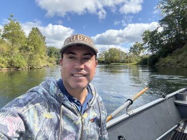 Selfie of an angler in a boat on a calm river, surrounded by lush greenery and a clear blue sky.