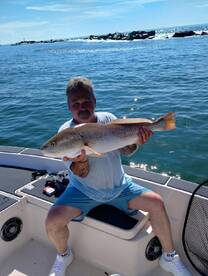 An angler proudly displaying a large Redfish while seated on a boat, with a scenic ocean backdrop.