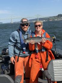 Two anglers proudly displaying a freshly caught Salmon while enjoying a day on the water near a scenic bridge.
