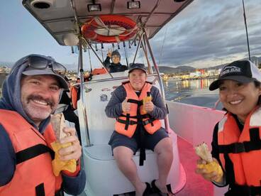 Group of anglers enjoying snacks on a fishing boat, wearing life jackets, with a scenic harbor view in the background.