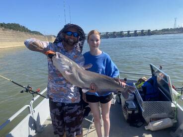 A young angler proudly displays a large Catfish alongside an adult on a fishing boat, enjoying a sunny day on the water.