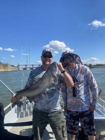 Two anglers proudly displaying a large Catfish while fishing on a boat, enjoying a sunny day on the water.
