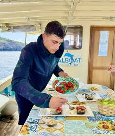 A young man preparing a meal on a boat, showcasing a vibrant dining setup with fresh ingredients and a scenic view of the coastline.
