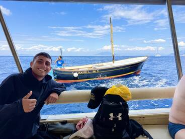 A young man enjoying a fishing trip, giving a thumbs-up while seated inside a boat, with another boat visible in the background on a sunny day.