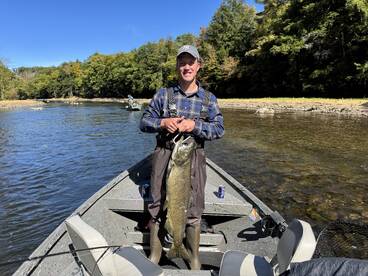 An angler proudly displaying a large fish while fishing on a river, surrounded by lush greenery and another boat in the background.