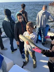 Two young anglers proudly displaying a striped bass while fishing on a boat, surrounded by fellow fishermen.