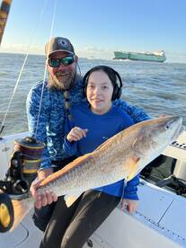 A young angler and an adult proudly displaying a large Redfish on a boat, enjoying a fishing trip in a coastal environment.