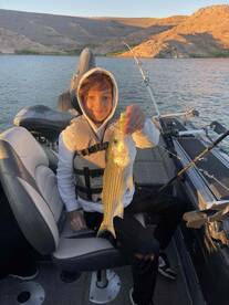 A young angler proudly displaying a striped bass while seated in a fishing boat, surrounded by a scenic landscape.