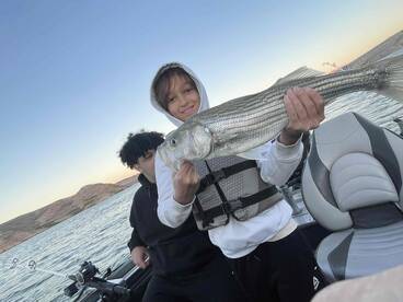 A young angler proudly displaying a striped bass while fishing on a boat, with scenic hills in the background.