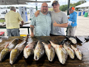 A proud duo poses with a variety of freshly caught fish on a display table at a fishing dock, showcasing their successful day on the water.