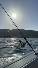 A fishing rod is poised over the shimmering water, capturing the essence of a sunny day on the water, with mountains in the background.