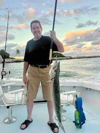 An angler proudly displaying a large King Mackerel aboard a fishing boat, with a scenic backdrop of clouds and palm trees.