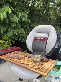 A wooden platter with grilled meats and dipping sauce is displayed on a boat seat, surrounded by lush greenery, highlighting a casual dining experience on the water.